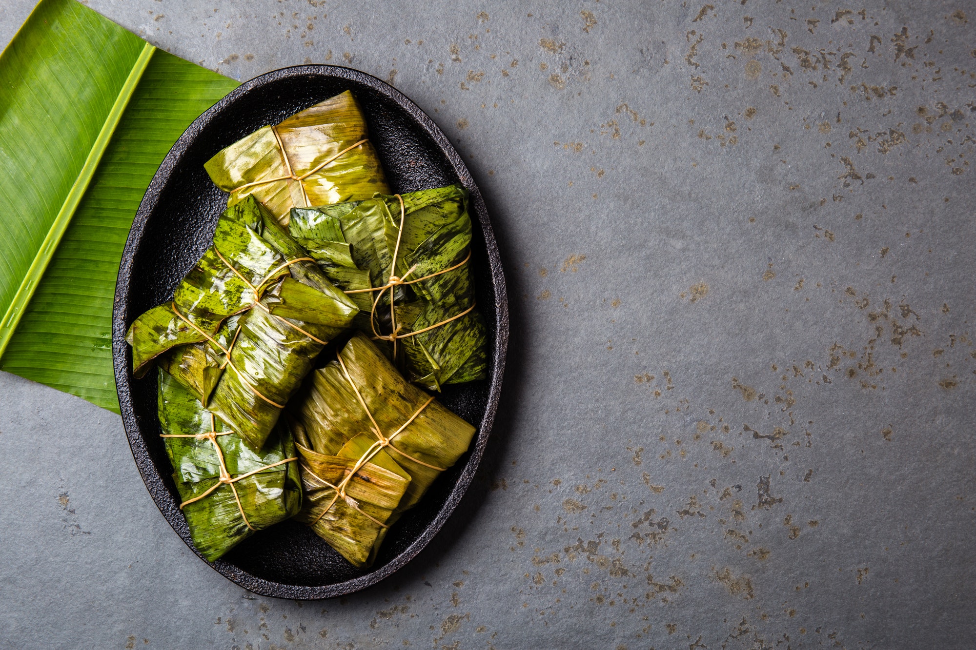 COLOMBIAN, CENTRAL AMERICAN FOOD. Tamales wrapped in banana palm tree leaves on black plate, gray